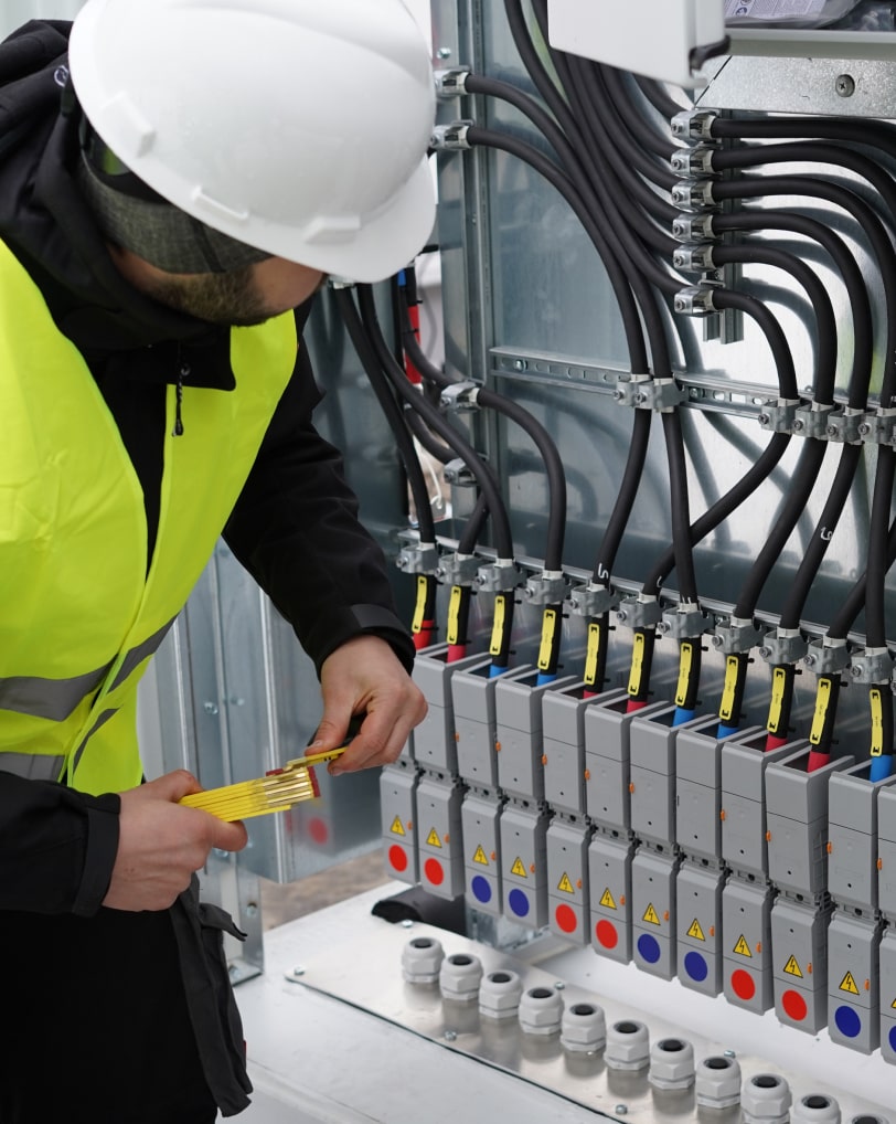 An employee wearing a hard hat and yellow safety vest stands in front of an open switch box, holding a folding rule.